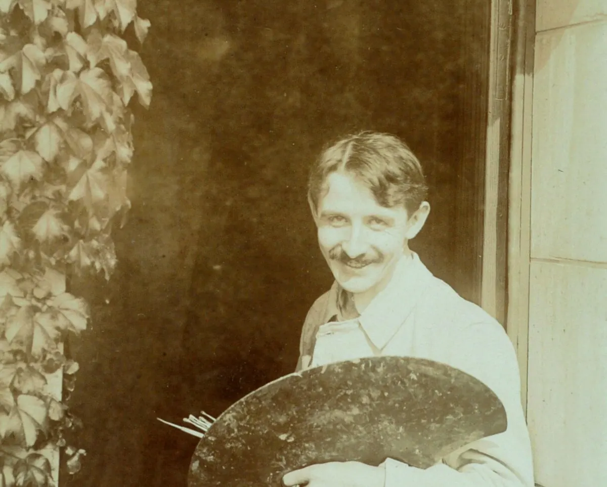 Jacques Majorelle 在常春藤覆蓋的拱門下、門口處微笑，手持畫家調色盤，約1910年的棕褐色照片